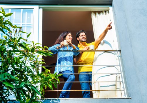 Young Indian couple enjoying morning coffee on balcony, sharing a relaxed conversation in warm sunlight, view from below